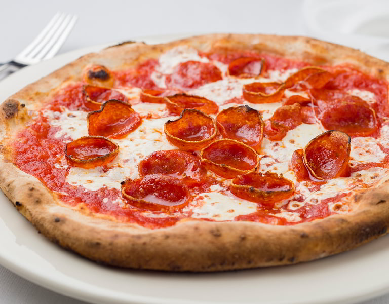 Pepperoni pizza with melted cheese and tomato sauce sits on a white plate beside a fork.