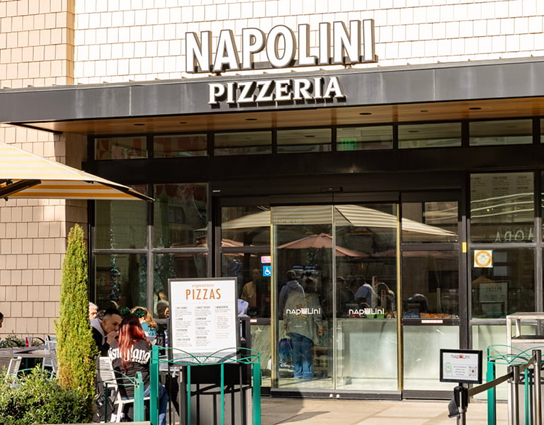 Napolini Pizzeria storefront with outdoor seating, menu signs, umbrellas, and glass entrance doors.
