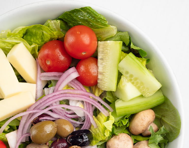 Green salad with tomatoes, cucumber slices, red onion, olives, mushrooms, and leafy greens in a white bowl.