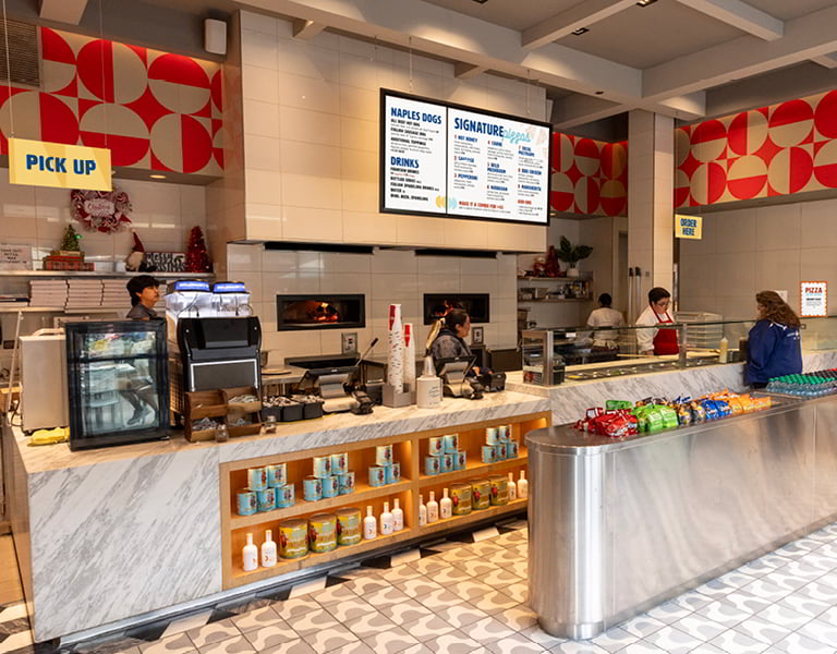 Restaurant counter with staff serving customers, pick up sign, overhead menus, packaged snacks, drinks, and a visible pizza oven.