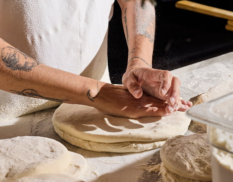 Hands press and shape pizza dough on a floured work surface surrounded by dough portions and a flour container.